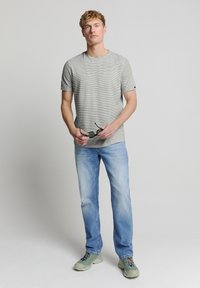 Grijs en wit gestreept T-shirt met ronde hals, gecombineerd met lichtblauwe jeans en lichtgroene schoenen. Model houdt een zonnebril vast, minimale details.