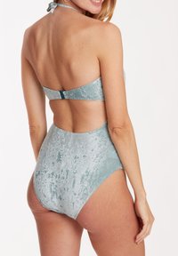Maillot de bain une pièce en tissu bleu clair texturé. Présente un col halter et une découpe dans le dos avec une sangle réglable et un fermoir.