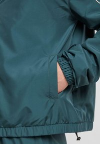 Teal vindjakke med elastiske mansjetter, sidelommer og samlet nederkant. Glatt, lett materiale med et uformelt design.