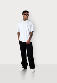 White short-sleeve t-shirt with a small, light-colored logo on the chest, paired with black wide-leg pants and white sneakers. Black sunglasses worn.