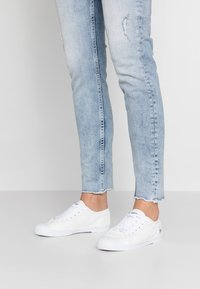 Baskets en toile blanches avec un motif texturé, dotées de bouts arrondis, d'un laçage sur le devant et de semelles en caoutchouc. Portées avec un jean déchiré bleu clair.