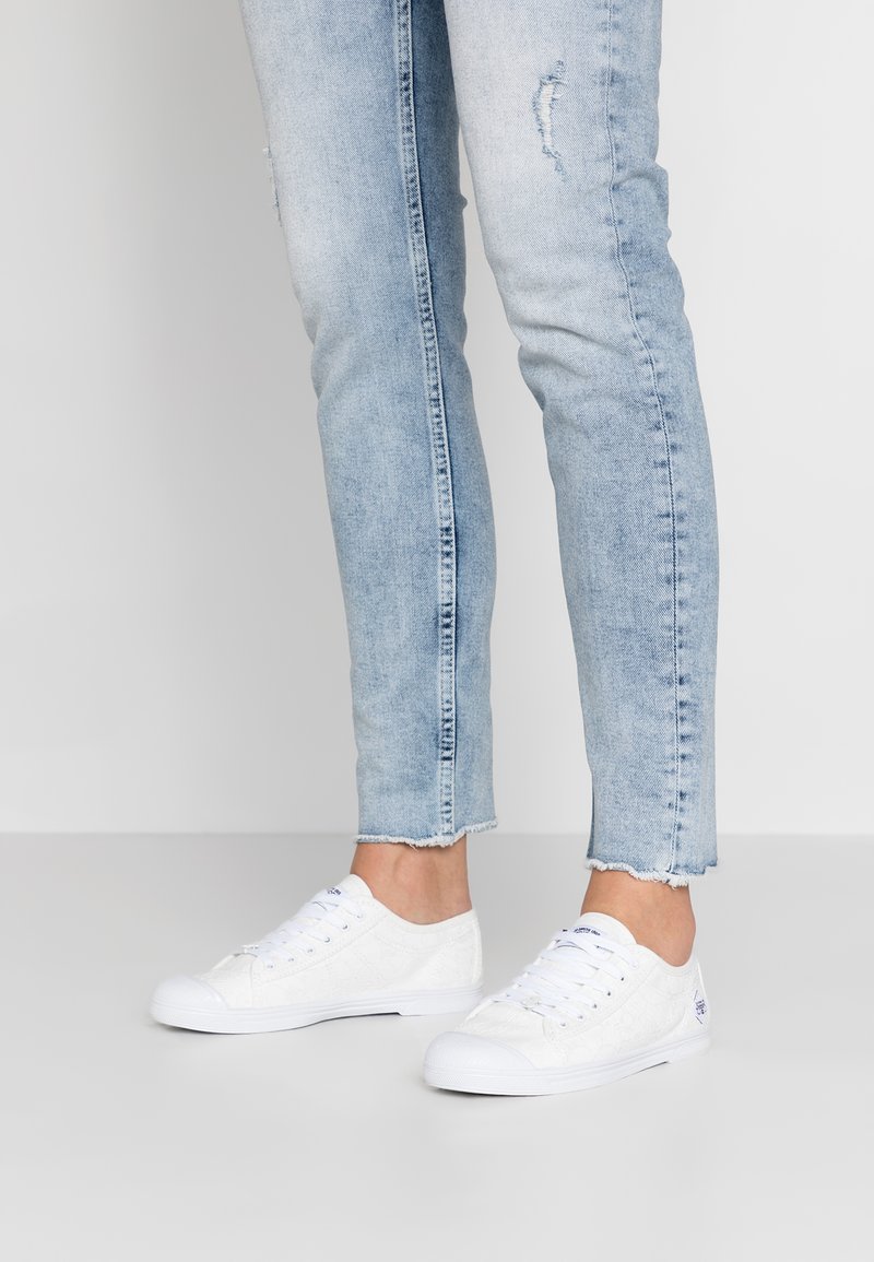 Baskets en toile blanches avec un motif texturé, dotées de bouts arrondis, d'un laçage sur le devant et de semelles en caoutchouc. Portées avec un jean déchiré bleu clair.