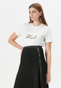 Modelo vistiendo una camiseta blanca con "Karl" en un bordado colorido, combinada con una falda negra brillante con botones dorados. Ella se presenta con confianza.