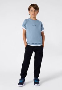 Zēns stāv ar rokām kabatās, ģērbies gaiši zilā īsām piedurknēm krekliņā, baltā apakškreklā, melnās biksēs un melni-zilās sporta kurpēs uz baltā fona.