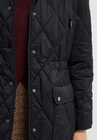 Czarna pikowana kurtka z zatrzaskami, posiada kieszeń z przodu, teksturowany materiał i regulowany detal przy szyi. Nosi się ją na bordowym swetrze.