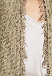 Gros plan d'une personne portant un cardigan en tricot beige texturé avec des franges par-dessus un haut blanc à encolure en V.