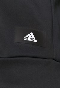 Gros plan sur un tissu noir avec une étiquette rectangulaire noire comportant le logo blanc Adidas et un texte.