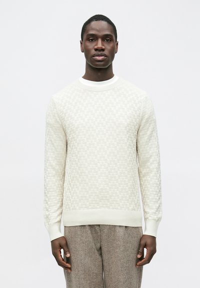 Pull beige clair avec un motif en damier texturé, col rond côtelé et manches longues, assorti à un pantalon gris.