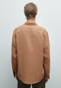 Chemise en lin marron avec une coupe droite, un ourlet arrondi, des poignets boutonnés et un col classique. Le tissu présente une apparence douce et texturée.