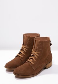 Bottines en daim marron avec lacets, dotées d'un petit talon carré, d'un bout arrondi et de perforations latérales pour la ventilation.