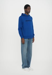 Uomo che indossa un felpa blu con la scritta "QUARTZ CO", jeans chiari e larghi, e scarpe da ginnastica bianche, in piedi contro uno sfondo grigio uniforme.