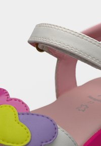 Παιδικό σανδάλι με λευκό λουράκι στον αστράγαλο, ροζ πάτο και διακοσμητικά λουλουδάκια σε ροζ, κίτρινο και μοβ χρώμα στην περιοχή των δαχτύλων.
