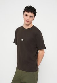 Καφέ βαμβακερό μπλουζάκι με κοντά μανίκια, στρογγυλή λαιμόκοψη και μικρό λευκό λογότυπο στο στήθος. Φορεμένο με πράσινο παντελόνι.