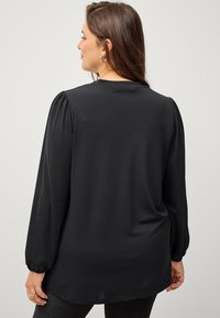 Blouse noire à manches longues en tissu doux et lisse, avec des détails froncés aux épaules, un col rond, une coupe ample et un ourlet incurvé.