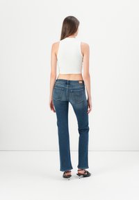 Sieviete stāv ar muguru pret skatītāju, ģērbusies baltā īsā topā, zilos taisnos džinsos un melnās papēžu kurpēs uz balta fona.