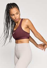 Bordeaux sportbh met een ronde hals en dikke bandjes, gecombineerd met hooggelegde lichtgrijze leggings. Glad materiaal met minimale stiksels.