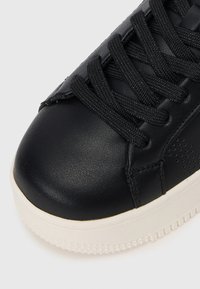 Black leather sneaker with textured finish, rounded toe, and flat black laces. Features a contrasting white rubber sole with grip pattern.