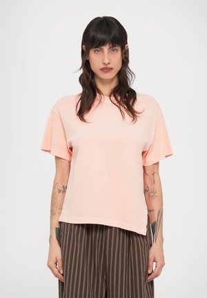 Ανοιχτό ροζ, υπερμεγέθη μπλουζάκι με κοντά μανίκια, στρογγυλή λαιμόκοψη και απαλή υφή. Συνδυασμένο με καφέ παντελόνι με κάθετες ρίγες.