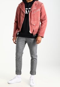 Persona che indossa una giacca motociclista in suede rosa, felpa nera con cappuccio, pantaloni grigi con risvolto, calzini bianchi e sneakers bianche, in piedi contro uno sfondo bianco.