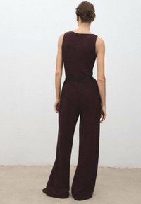 Burgundy jumpsuit med figursydd liv och vida ben, i texturerat tyg, samt ett svart bälte i midjan, sedd bakifrån.