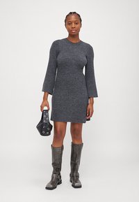 Robe en tricot côtelé gris avec de longues manches évasées, portée avec des bottes gris jusqu'au genou et un petit sac noir orné d'accents circulaires.
