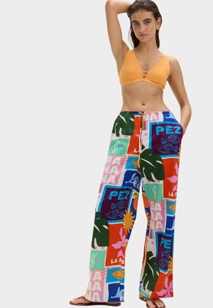 Kobieta ubrana w pomarańczowy top od bikini i kolorowe spodnie o szerokich nogawkach z tropikalnymi i tekstowymi wzorami, stoi z jedną uniesioną ręką i jedną ręką w kieszeni.