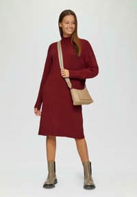 Burgunderfarbenes Strickkleid mit hohem Kragen und langen Ärmeln, kombiniert mit einer beigen Umhängetasche und grauen Knöchelstiefeln. Glatte Textur, knielang.