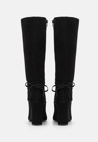 Botas de ante negras hasta la rodilla con un tacón grueso, que cuentan con un detalle de lazo en la parte trasera y un cierre de cremallera para facilitar su uso.