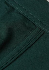 Tmavě zelená tkanina s hladkým povrchem, zdobená vrstvenými záhyby a prošívaným lemem. Zblízka se zaměřuje na vazbu a strukturu látky.