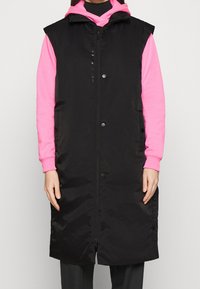 Svart vadderad väst med dragkedja framtill och tryckknappar, med hög krage och ärmlös design. Kombinerad med en rosa huvtröja under.