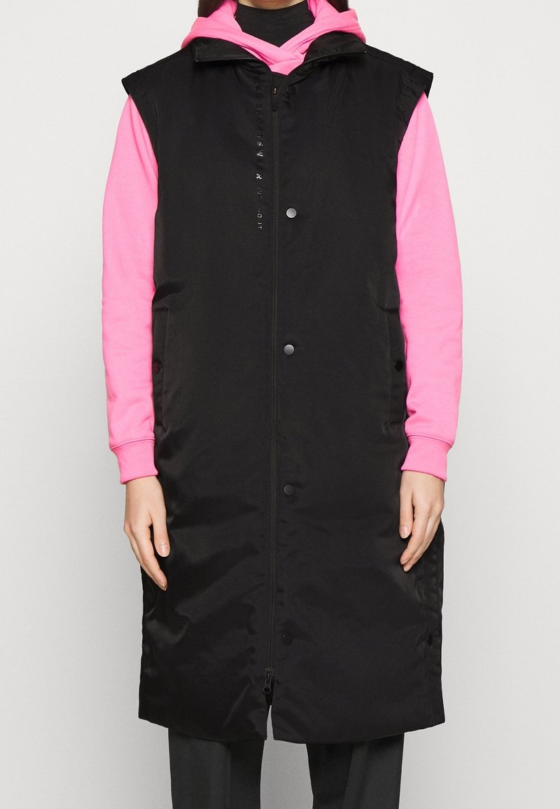 Svart vadderad väst med dragkedja framtill och tryckknappar, med hög krage och ärmlös design. Kombinerad med en rosa huvtröja under.