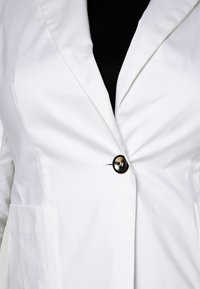 Bílý blazer vyrobený z texturované tkaniny, s klopovým límcem, jedním černým knoflíkem na zapínání a dvěma předními kapsami.