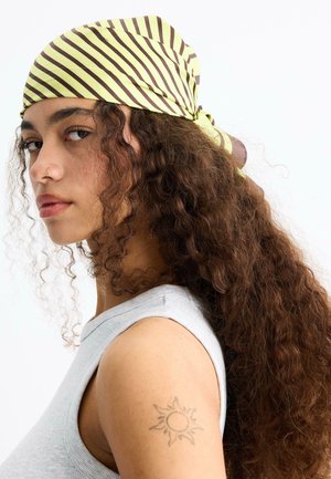 Mladá žena s dlhými kučeravými vlasmi, ktorá nosí žlto-hnedý pruhovaný šál na hlave, sivý tielkový top, krúžok v nose a tetovanie slnka na hornej časti ramena.