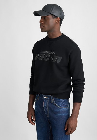 Schwarzer Pullover mit erhabenem "DSQUARED2 DUCATI" Schriftzug, kombiniert mit blauen Jeans. Das Model trägt eine schwarze Lederkappe.
