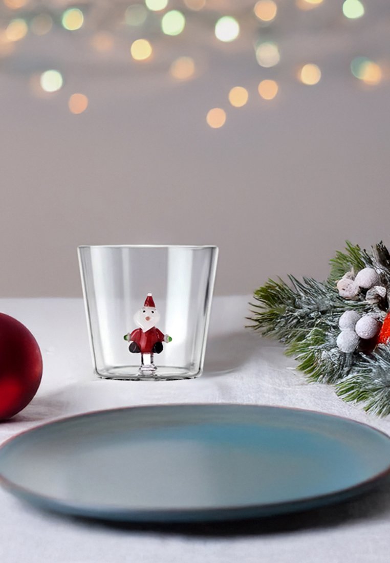 Bicchiere di vetro con una figura di Babbo Natale all'interno, con accenti rossi e bianchi. Le caratteristiche circostanti includono un ornamento rosso, un piatto blu e fogliame verde.