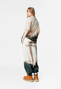 Camiseta gráfica de gran tamaño en tonos beige y verde con textura, combinada con pantalones holgados que exhiben un estampado de paisaje y letras en negrita. Botas marrones.