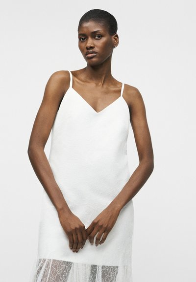 Γυναίκα με σκουρόχρωμο δέρμα φοράει λευκό αμάνικο φόρεμα με δαντελωτό τελείωμα, στέκεται μπροστά σε απλό ανοιχτόχρωμο φόντο, τα χέρια της χαλαρά συγκρατημένα.