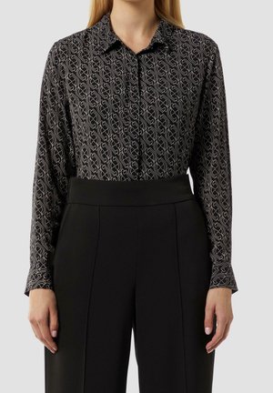 Femme portant une chemise noire à manches longues avec des motifs géométriques blancs et un pantalon noir taille haute sur un fond uni.