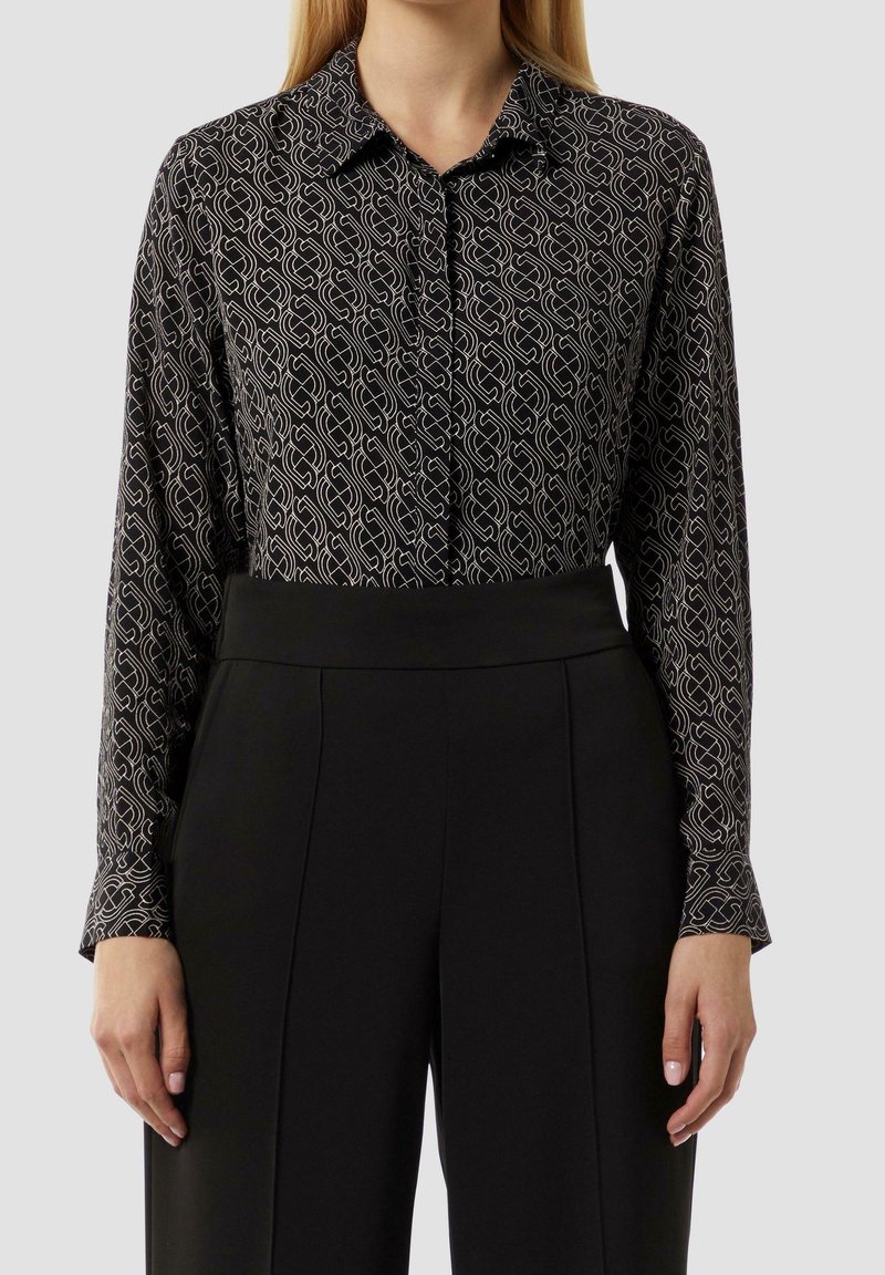 Femme portant une chemise noire à manches longues avec des motifs géométriques blancs et un pantalon noir taille haute sur un fond uni.