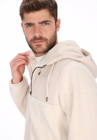 Sudadera con capucha de forro polar beige, con un frente de tela suave, cuerpo texturizado y cordón ajustable. Cuenta con cremallera frontal y bolsillos laterales.