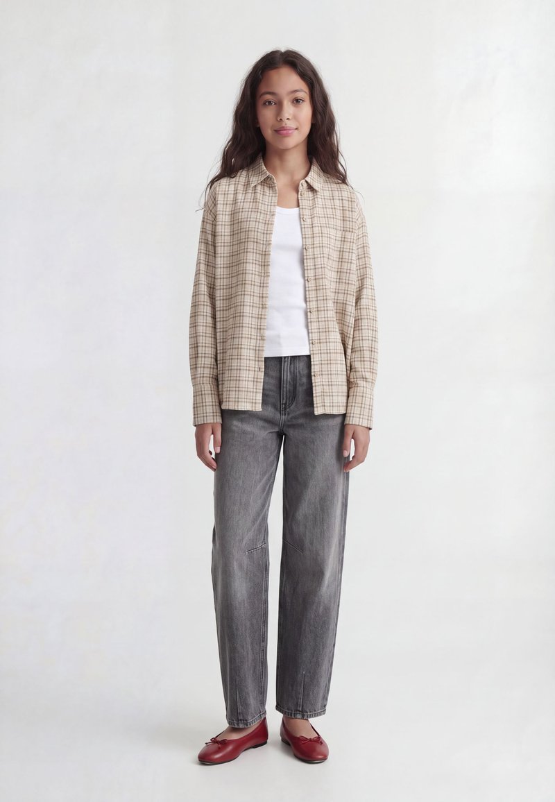 Jeune femme debout, portant une chemise à carreaux beige sur un haut blanc, jean gris et ballerines rouges sur un fond clair uni.
