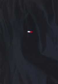 Czarny materiał o gładkiej fakturze z małym haftowanym logo w kolorach czerwonym, białym i niebieskim, umiejscowionym po lewej stronie.