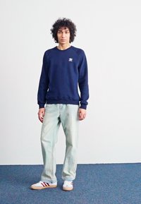 Marineblå sweatshirt med rund halsudskæring og hvidt logo på venstre bryst, kombineret med lyseblå denimjeans og hvide sneakers med røde striber.