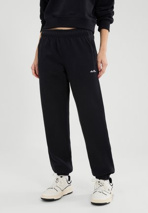 Person trägt schwarze Ellesse-Sweathosen, schwarzen kurz geschnittenen Pullover und weiße Sneaker mit schwarzen Details, steht vor einem weißen Hintergrund.