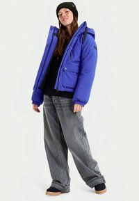 Jachetă puffer albastră cu glugă, mansete elastice și multiple buzunare, purtată cu blugi largi gri și bocanci negri.