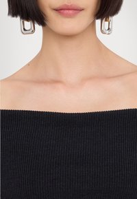 Femme portant de grandes boucles d'oreilles rectangulaires argentées et un haut noir côtelé épaules dénudées, avec des cheveux courts et foncés visibles sur les bords.