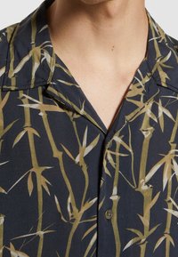 Camicia a maniche corte con un tessuto scuro e una stampa di bambù in tonalità di verde e beige. Include un colletto a V e una chiusura con bottoni.