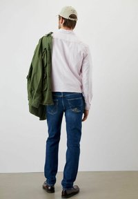 Giacca verde drappeggiata sulla spalla, camicia rosa, jeans blu con design distintivo sulla tasca posteriore e scarpe marroni. Sfondo neutro.