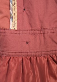 Roestkleurige jurk met een geplooide onderkant, gemaakt van een textuur stof en een decoratieve, gemusterde afwerking langs één kant.