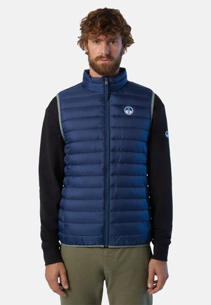 Gilet in piuma blu navy con cuciture orizzontali, colletto alto e logo sul petto. Camicia nera a maniche lunghe sotto con accenti a contrasto.